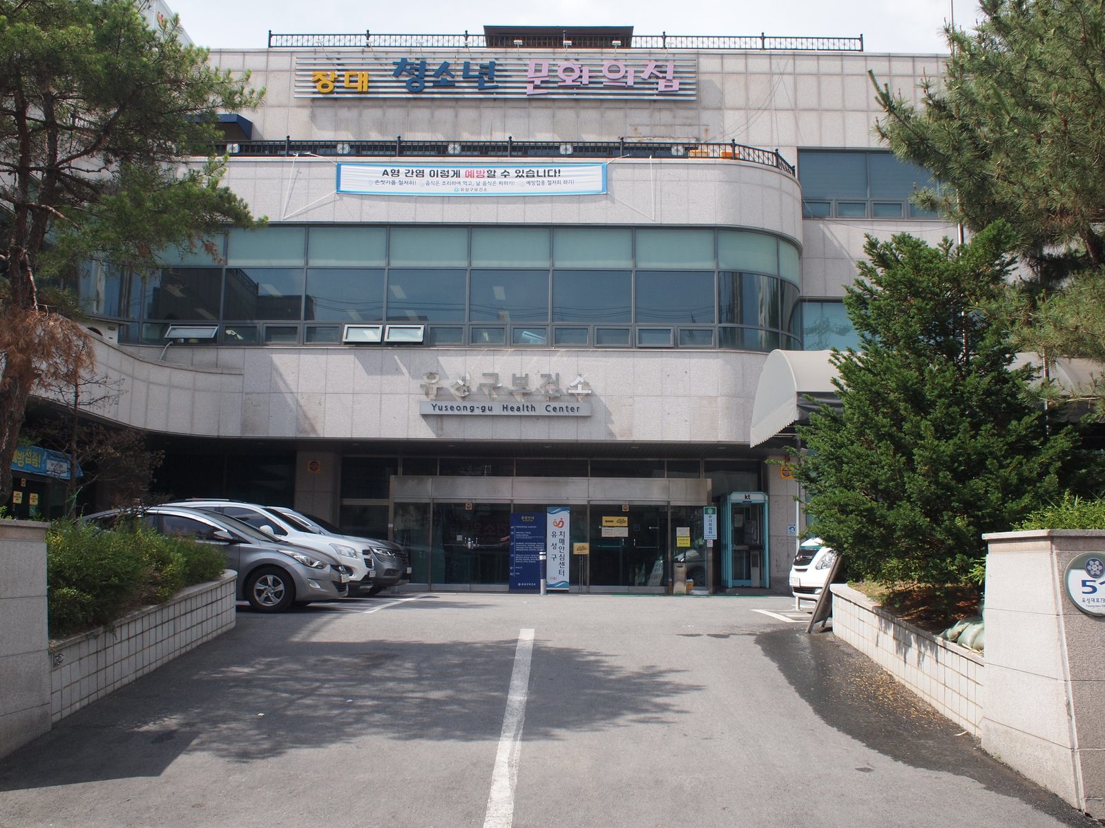 '유성구보건소'라고 돌벽에 적힌 큰 건물이에요. 이층은 유리가 둥글게 둘러져 있어요. 건물 앞 주차장에 자동차가 서 있어요.