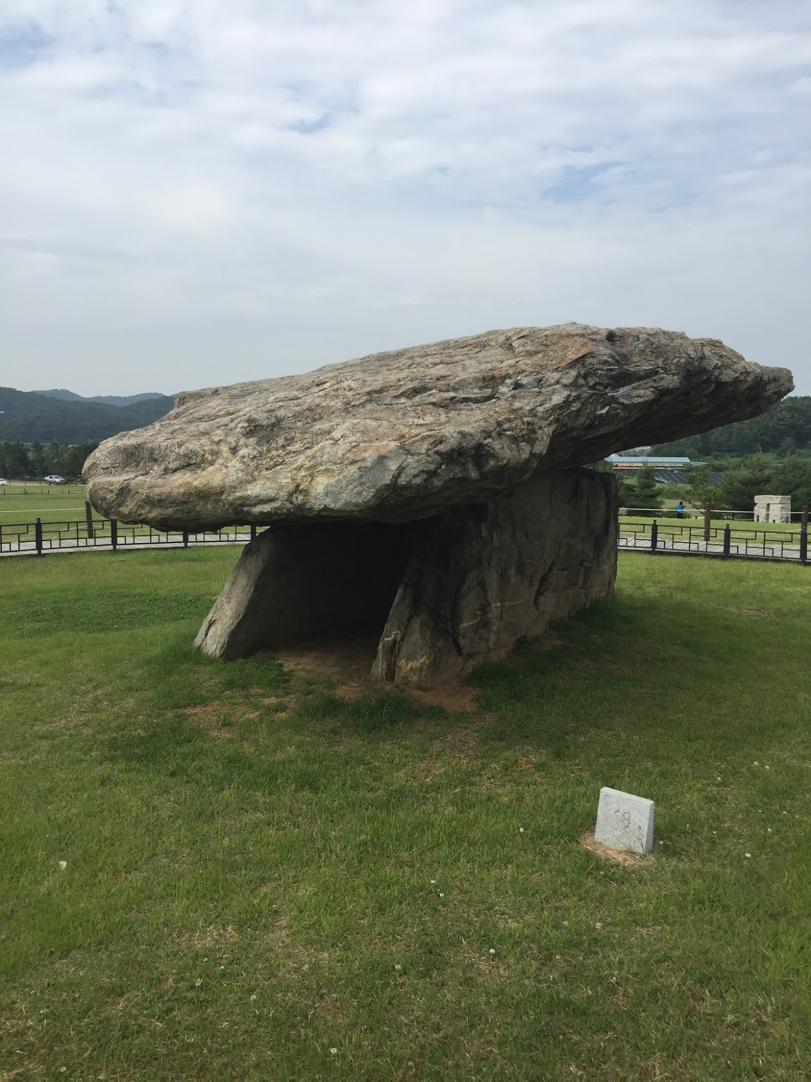 넓은 잔디밭 가운데에 커다랗고 넓적한 돌이 두 개의 받침돌 위에 탁자처럼 얹혀 있다. 앞에 작은 흰 표지석이 있고 뒤로 낮은 울타리와 산이 보인다.