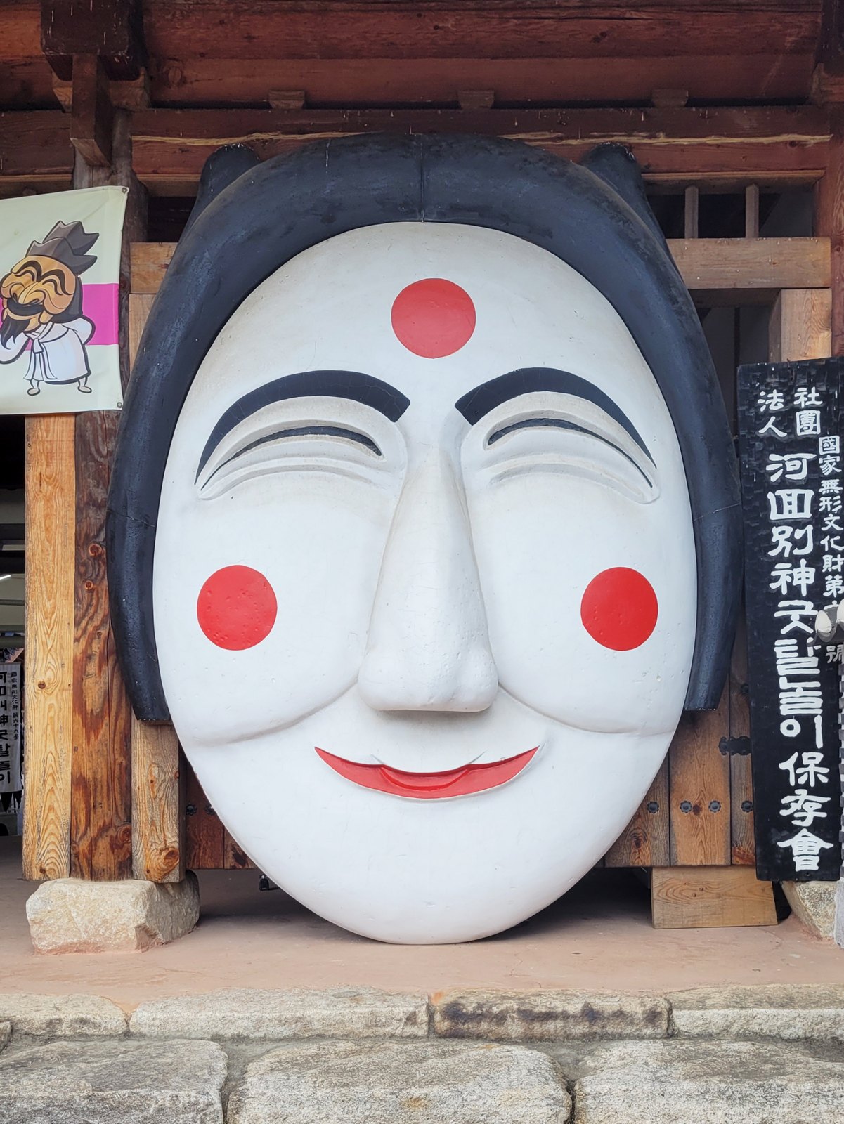 나무 건물 기둥 사이에 사람 키보다 큰 하얀 탈이 서 있다. 이마와 양 볼에 빨간 동그라미, 검은 눈썹, 웃는 입. 오른쪽에 검은 세로 간판이 있다.