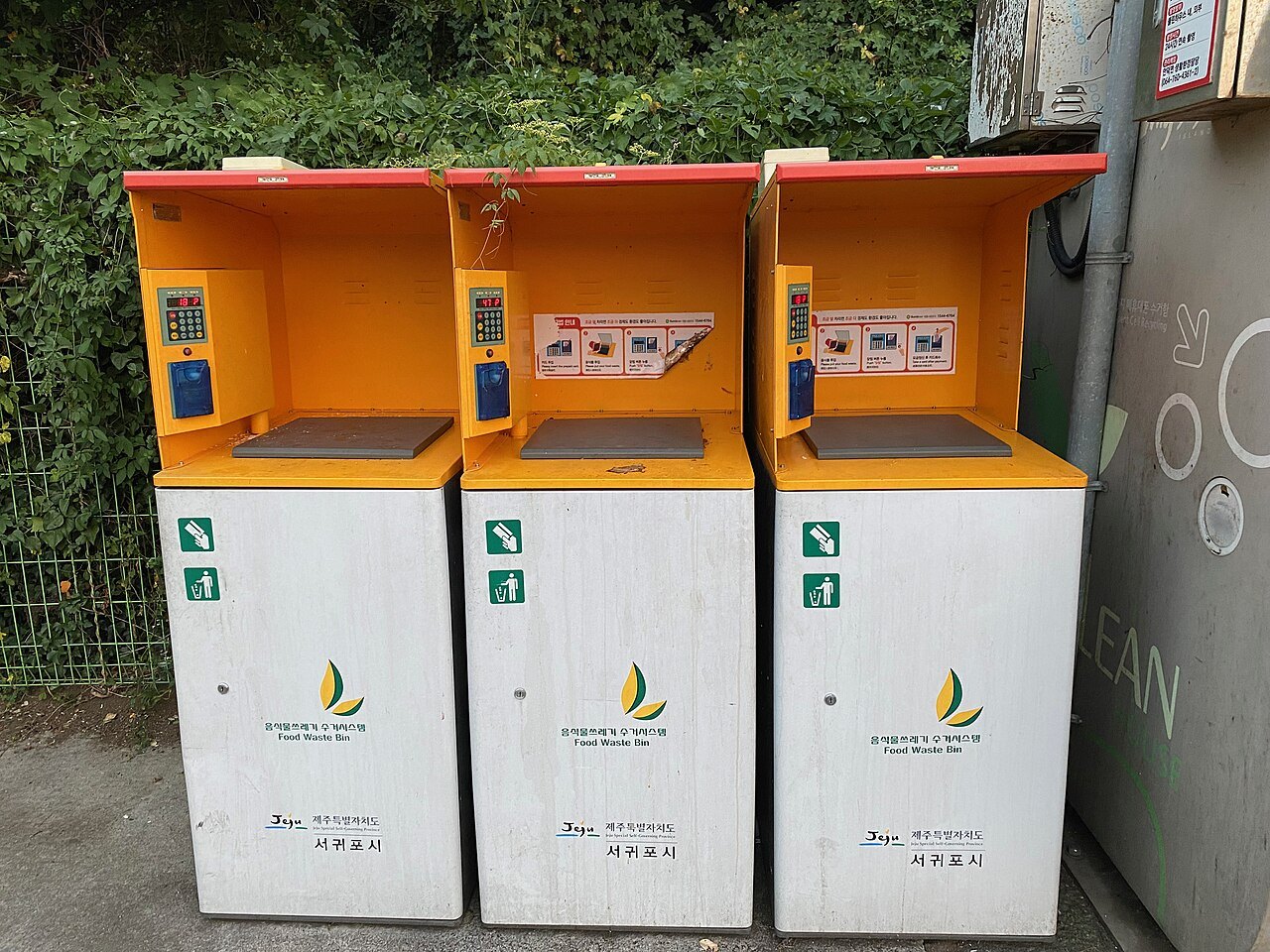 수풀 앞에 나란히 선 음식물쓰레기 수거함 세 대. 주황색 윗부분에 숫자 표시창과 버튼, 파란 카드 인식기가 달려 있고, 흰 몸체에 '음식물쓰레기 수거시스템 Food Waste Bin', '제주특별자치도 서귀포시'라고 적혀 있다
