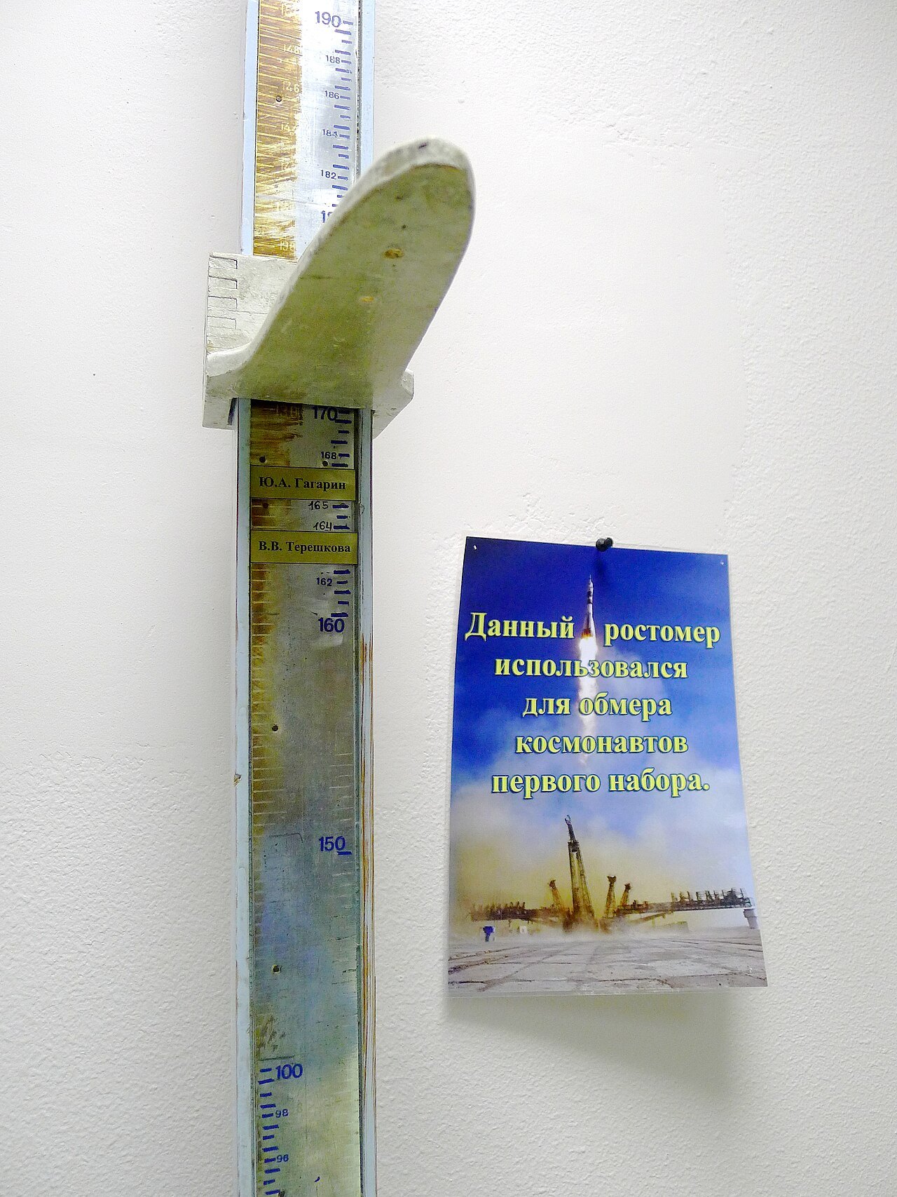 흰 벽에 세워진 오래된 신장계. 센티미터 눈금 막대에 머리판이 걸려 있고, 160과 170 사이에 가가린과 테레시코바의 이름판이 붙어 있으며, 옆에 러시아어 안내문이 걸려 있다
