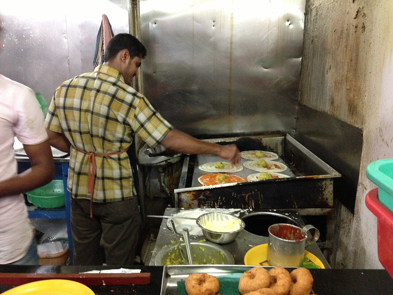 좁은 식당 주방의 넓은 철판에서 도사(인도식 부침) 여러 장을 한꺼번에 굽는 요리사. 앞 진열대에는 도넛 모양의 튀김이 놓여 있다
