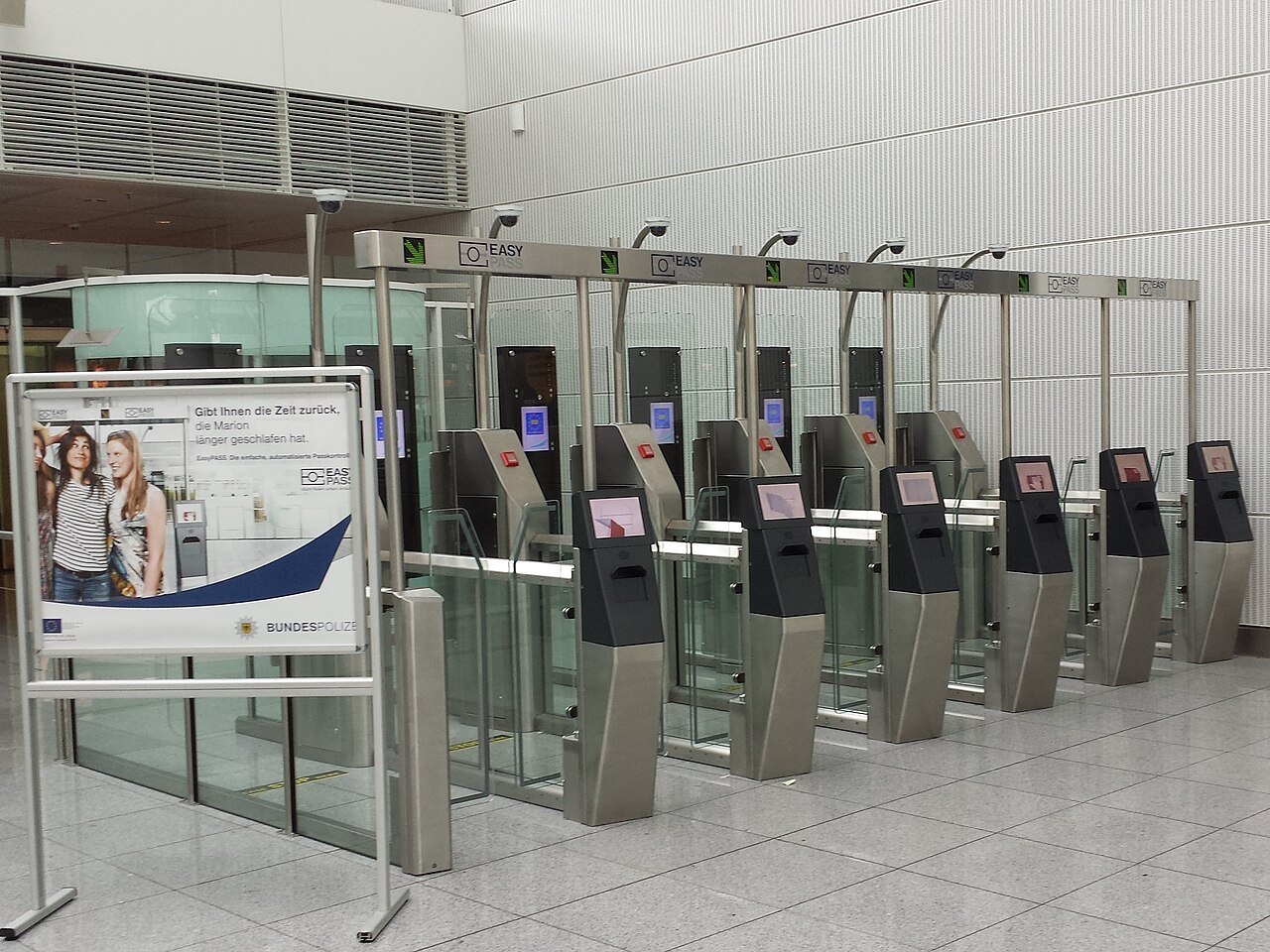 공항 안에 나란히 선 자동 출입국 심사대 여섯 대. 기둥마다 카메라가 달려 있고 여권을 올리는 단말과 유리문이 있으며, 왼쪽에 안내 광고판이 서 있다